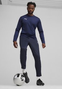 Marinefarbendes, sportliches Langarmshirt und enganliegende Hosen aus synthetischem Material, kombiniert mit schwarzen Fußballschuhen und einem gemusterten Fußball.
