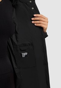 Hand die een open zwarte, gequiltte jas vasthoudt met een binnenzak waar het label "Khujo TM original for a few" op staat, tegen een donkere kledingachtergrond.