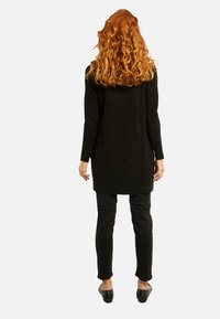 Černá žebrovaná tunika s dlouhým rukávem a kulatým spodním lemem, spárovaná s černými přiléhavými kalhotami a černými plochými botami. Vlnité vlasy sahající po ramena.