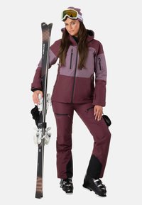 Completo da sci in bordeaux e lilla, composto da giacca con cappuccio e pantaloni con cerniere, abbinato a sci e scarponi da sci.