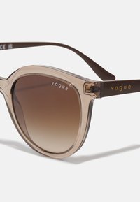 Ochelari de soare cu un cadru transparent de culoare maro, lentile maronii gradient și branding auriu "vogue" pe tâmple. Formă rotundă cu detalii de design elegante.