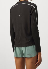 Černé dlouhé rukávy sportovní tričko s reflexními bílými pruhy, strukturovaná tkanina a kulatým detailem na zádech. Kombinováno s mintově zelenými šortkami.