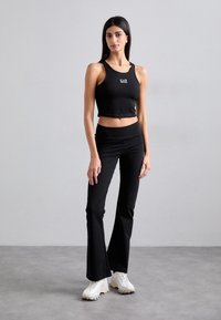 Črn croptop z logotipom je vparen z visokimi črnimi širokimi hlačami. Model nosi bele superge. Gladek, oprijeten material, minimalen dizajn.
