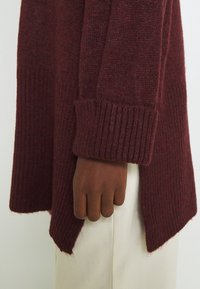 Maglione bordeaux in maglia con polsini e orlo a coste. Presenta una vestibilità comoda, spacchi laterali e una texture morbida, abbinato a pantaloni di colore chiaro.