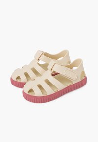 Light beige rubber sandals with open sides, adjustable strap, and textured pink sole. Features cut-outs for ventilation and thumb grip accents.