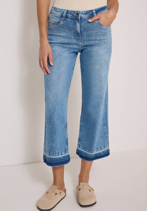 Femme portant un jean flare court bleu clair avec ourlets retroussés et des chaussures beiges sans lacets, debout devant un fond uni.