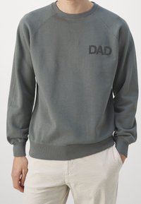 Muž v šedé mikině s textem "DAD" na hrudi a světlých kalhotách, s jednou rukou v kapse na jednobarevném pozadí.