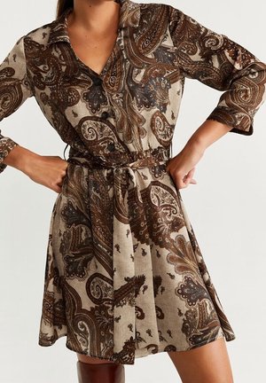 Vrouw die een bruine jurk met paisley patroon draagt, met knopen, driekwart mouwen, een stoffen tailleband en kniehoge bruine laarzen.