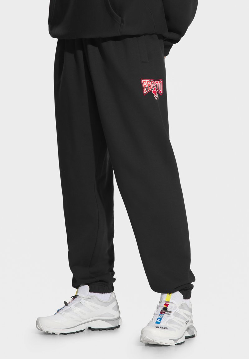 Czarne dresy z czerwonym logo "Presto" na lewym udzie, z elastycznym paskiem w talii i mankietami, połączone z białymi butami sportowymi.
