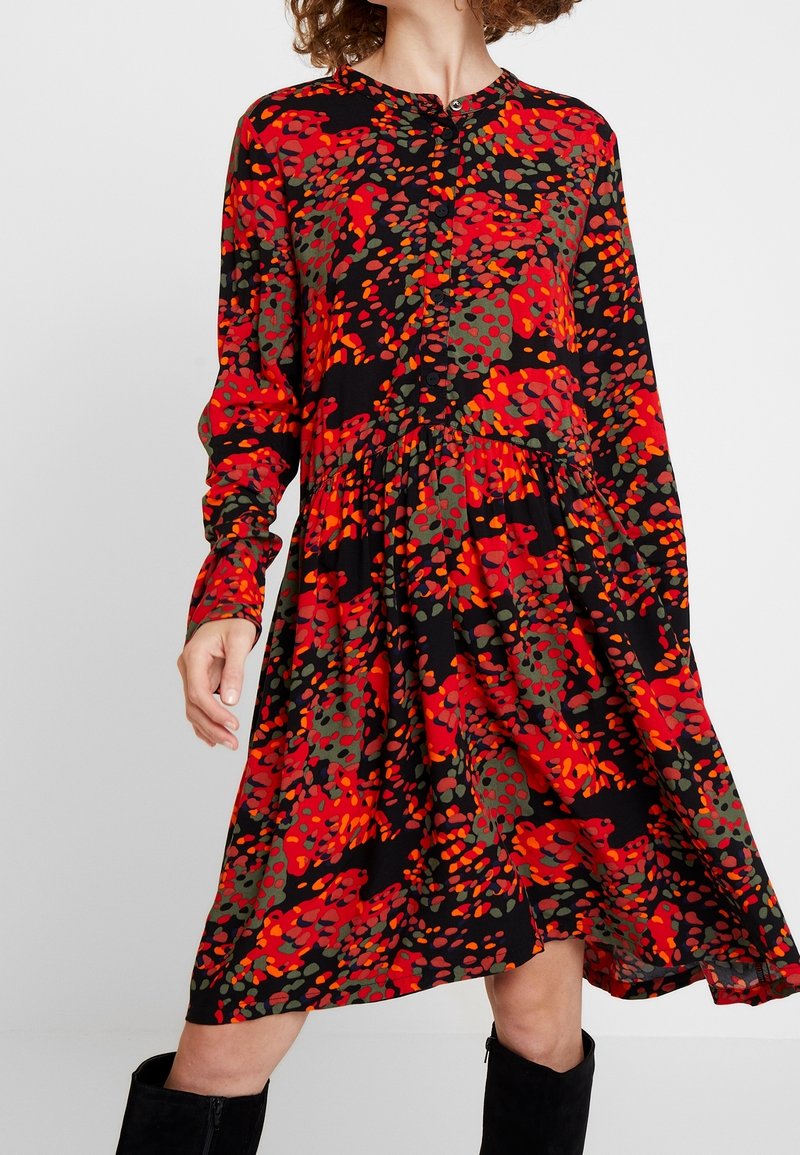 Femme portant une robe à manches longues avec un motif abstrait rouge, orange et vert, associée à des bottes noires montantes.