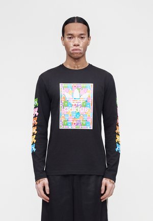 Person mit Vitiligo, die ein schwarzes Langarmshirt trägt, das bunte Teddybären und ein mehrfarbiges Adidas-Logo auf Vorderseite und Ärmeln zeigt.