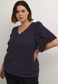 Lilla struktureret top med V-hals, korte puffærmer og afslappet pasform. Stoffet har et subtilt geometrisk mønster. Bæres med sorte bukser.