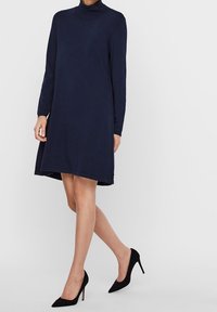 Kvinna iklädd en marinblå långärmad polokragsklänning kombinerad med svarta spetsiga högklackade skor, stående mot en enkel ljus bakgrund.