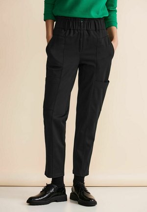 Schwarze tapered Hose mit einem elastischen Bund, zwei Seitentaschen und sichtbaren Nähten. Kombiniert mit schwarzen Schuhen und einem grünen Oberteil.