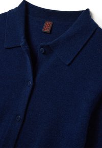 Cardigan en maille bleu marine avec un col, trois boutons de fermeture et une finition douce et texturée. Étiquette visible avec le nom de la marque.