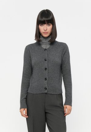 Grauer Strickcardigan mit rundem Halsausschnitt, gerippter Säume und sechs schwarzen Knöpfen. Wird über einem grauen Rollkragenpullover getragen. Weicher Stoff und figurbetontes Design.
