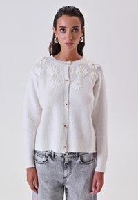 Cardigan bianco lavorato a maglia con applicazioni floreali sulle spalle, sei bottoni nella parte anteriore e polsini e orlo a coste.