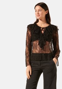 Blouse en dentelle noire avec un col froncé, de longues manches transparentes et un détail à nouer au cou, ainsi qu'une taille en dentelle ajustée.
