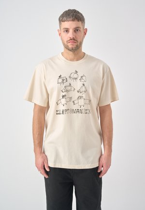 Man met kort haar en oorbellen draagt een crèmekleurig T-shirt met abstracte vogeltekeningen en tekst, staand tegen een effen witte achtergrond.