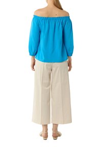 Niebieska bluzka na jedno ramię z bufiastymi rękawami, połączona z kremowymi spodniami o szerokich nogawkach i beżowymi sandałami. Gładki materiał z elastycznym dekoltem.
