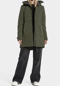 Parka impermeable verde oliva, con cremallera frontal, capucha con cordón y dos grandes bolsillos en el pecho. Usada sobre jeans de pierna ancha negros y zapatillas blancas.