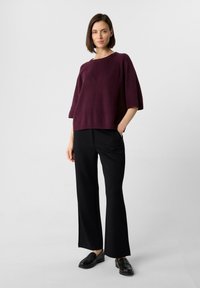 Burgunderfarbener Strickpullover mit lockerer Passform und kurzen Ärmeln, kombiniert mit schwarzen weit geschnittenen Hosen und schwarzen Slippern.