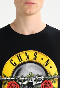 Zwarte katoenen t-shirt met een cirkelvormige gele grafiek van een schedel, een geweer en rode rozen. Ronde halslijn en korte mouwen.