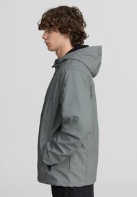 Giacca a hood grigia con una finitura liscia e opaca, dotata di cerniera frontale, tasche laterali e fondo regolabile. Indossata da un individuo di lato.