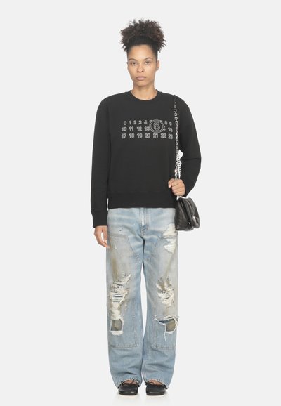 Felpa nera con numeri grafici, jeans azzurri chiari consumati, borsa nera con tracolla a catena e scarpe nere. Tessuto morbido, vestibilità rilassata.