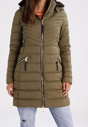 Veste longue matelassée gonflée vert olive avec fermeture éclair frontale et poches zippées en biais, portée sur un jean bleu clair déchiré.
