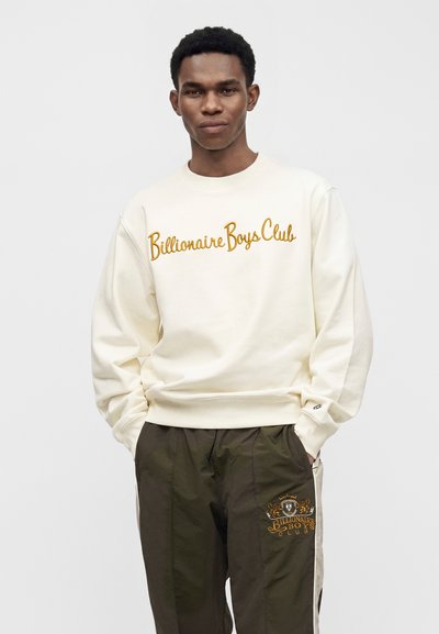 Άνδρας που φοράει κρεμ φούτερ με τη φράση «Billionaire Boys Club» και σκούρο παντελόνι με το ίδιο λογότυπο, στέκεται με τα χέρια στις τσέπες.