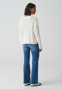 Maglione bianco a maglia texturizzata con una vestibilità ampia, abbinato a jeans blu a zampa. Il maglione presenta un collo rotondo e dettagli a coste.