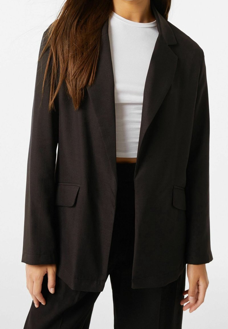 Femme portant un blazer noir et un pantalon avec un top court blanc, debout devant un fond blanc.