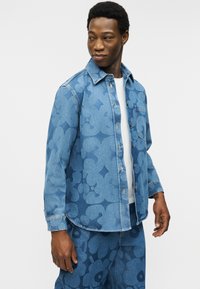 Νεαρός άνδρας που φοράει μπλε τζιν μπουφάν με λουλουδάτο σχέδιο και παντελόνι που ταιριάζει, κοιτάζοντας πλάγια με ελαφρύ χαμόγελο.