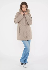 Cappotto in beige per l'inverno con colletto rifinito in pelliccia, caratterizzato da un taglio dritto, tasche laterali, indossato sopra jeans svasati blu e sneakers bianche.