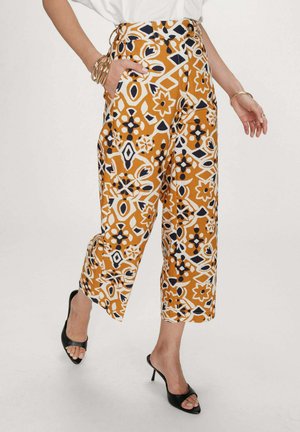 Femme portant un pantalon court à jambes larges avec des motifs géométriques floraux orange, noir et blanc, des talons ouverts noirs et des bracelets dorés.