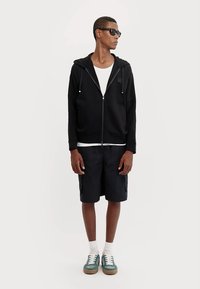 Sudadera negra con cremallera y cordones, camiseta blanca, pantalones cortos oscuros y zapatillas verdes con suelas marrones. El modelo lleva gafas de sol.