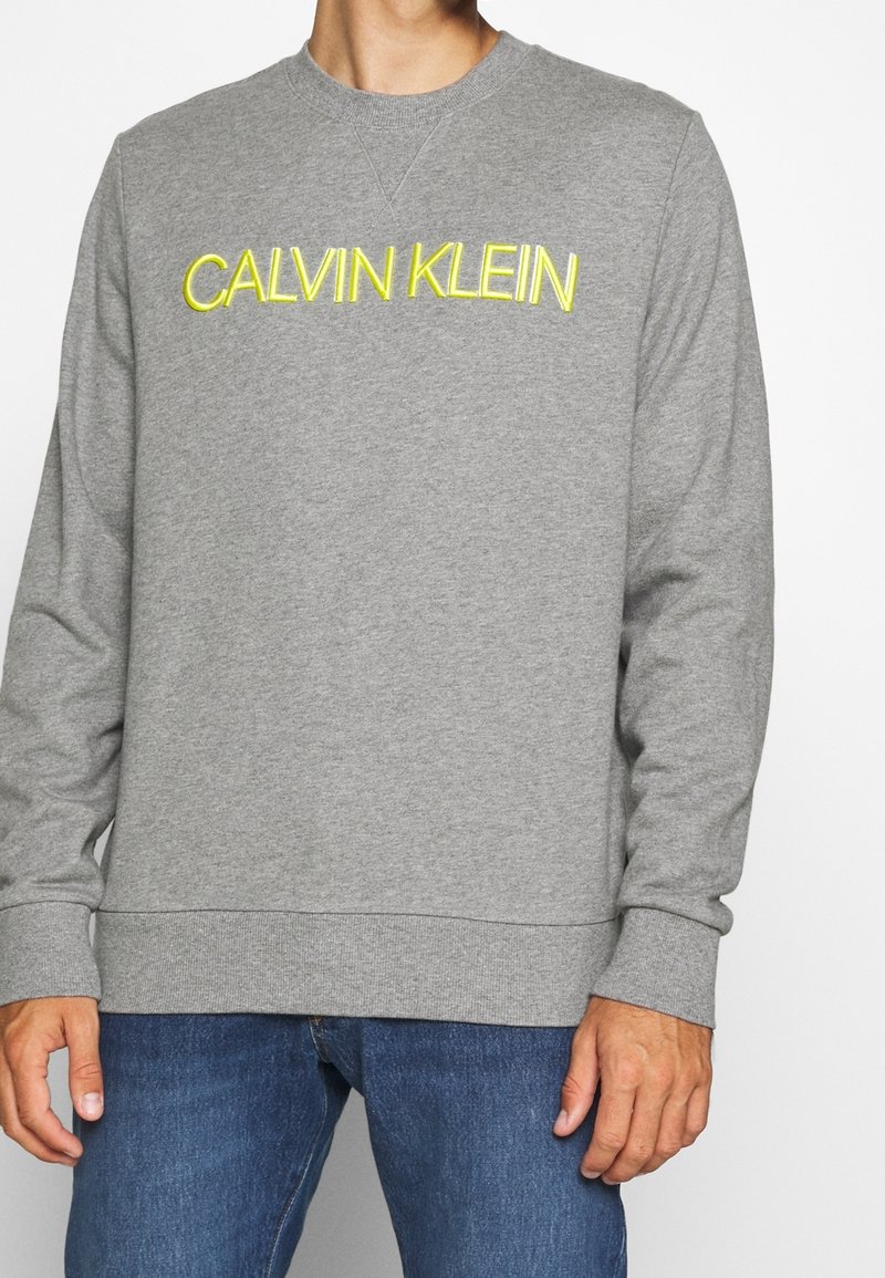 Sweat-shirt gris avec broderie jaune « CALVIN KLEIN », poignets et ourlet côtelés, et design classique à col rond.