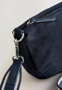 Esquina de bolso de ante azul oscuro con cremallera plateada, cierre metálico y correa de tela a rayas azul marino unida a un anillo de metal.