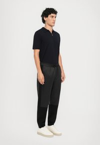 Svart polo skjorte med glidelås og svarte bukser med elastisk midje, som har teksturerte sidepaneler, kombinert med hvite sko. Nøytral bakgrunn.