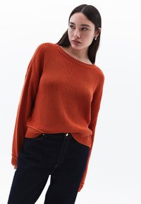 Trui in oranje ribgebreide stof, brede bootnek, oversized fit, lange mouwen, textuurdetail. Gepaard met donkerblauwe jeans.