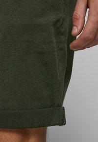 Tmavě zelené šortky vyrobené z texturované látky, s ohrnutým lemem a detaily bočních švů. Detailní pohled na materiál a design.