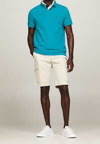 Turkusowa koszulka polo z krótkim rękawem i kołnierzykiem, w zestawie z beżowymi szortami cargo z bocznymi kieszeniami oraz białymi sneakersami.