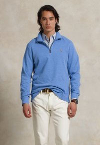Sweater de pullover azul claro com zíper em tecido suave, combinado com calças brancas, apresentando um pequeno logotipo no peito e sobreposto a uma camisa com colarinho.