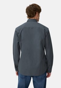 Man in een donkergrijze lange mouwen overhemd met knopen en blauwe spijkerbroek, gezien van achteren tegen een effen achtergrond.