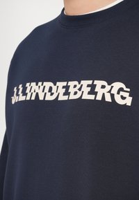 Σκούρο μπλε φούτερ με στρογγυλή λαιμόκοψη, με μεγάλο λευκό κείμενο "J.LINDEBERG". Μαλακή υφή και μινιμαλιστικός σχεδιασμός, κοντινή λήψη στην περιοχή του στήθους.