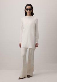 Sieviete, tērpusies krēmkrāsas garās piedurkņu tunikā, atbilstošās flared biksēs, brūnās šaurās kurpēs, melnās saulesbrillēs un krēmkrāsas auskaros, stāv pret vienkāršu fona.