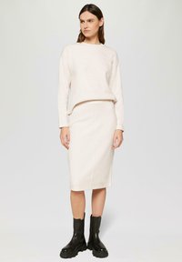 Hellbeigees Strickpullover, kombiniert mit einem passenden Midirock, der eine schlichte Silhouette präsentiert. Schwarze, klobige Stiefeletten runden das Outfit ab.
