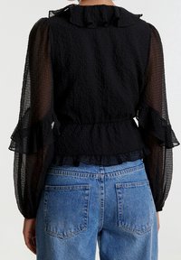 Blouse noire en tissu texturé, col à volants, manches longues transparentes, associée à un jean en denim bleu taille haute.