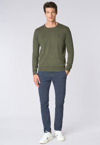 Maglione a maglia verde oliva con scollo a girocollo, abbinato a pantaloni blu navy e sneakers bianche, con un logo discreto sul petto.